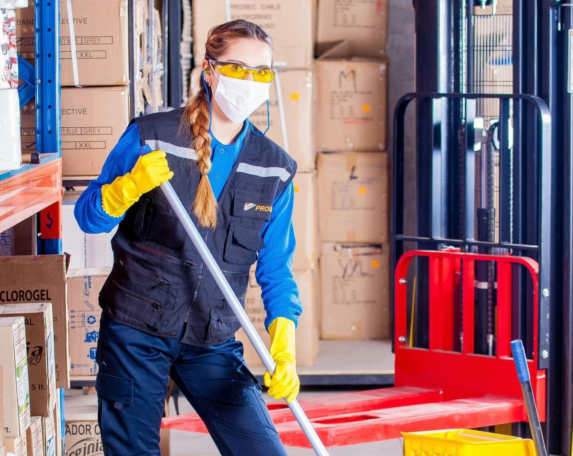 A woman wearing a mask and gloves is mopping the floor in a warehouse.