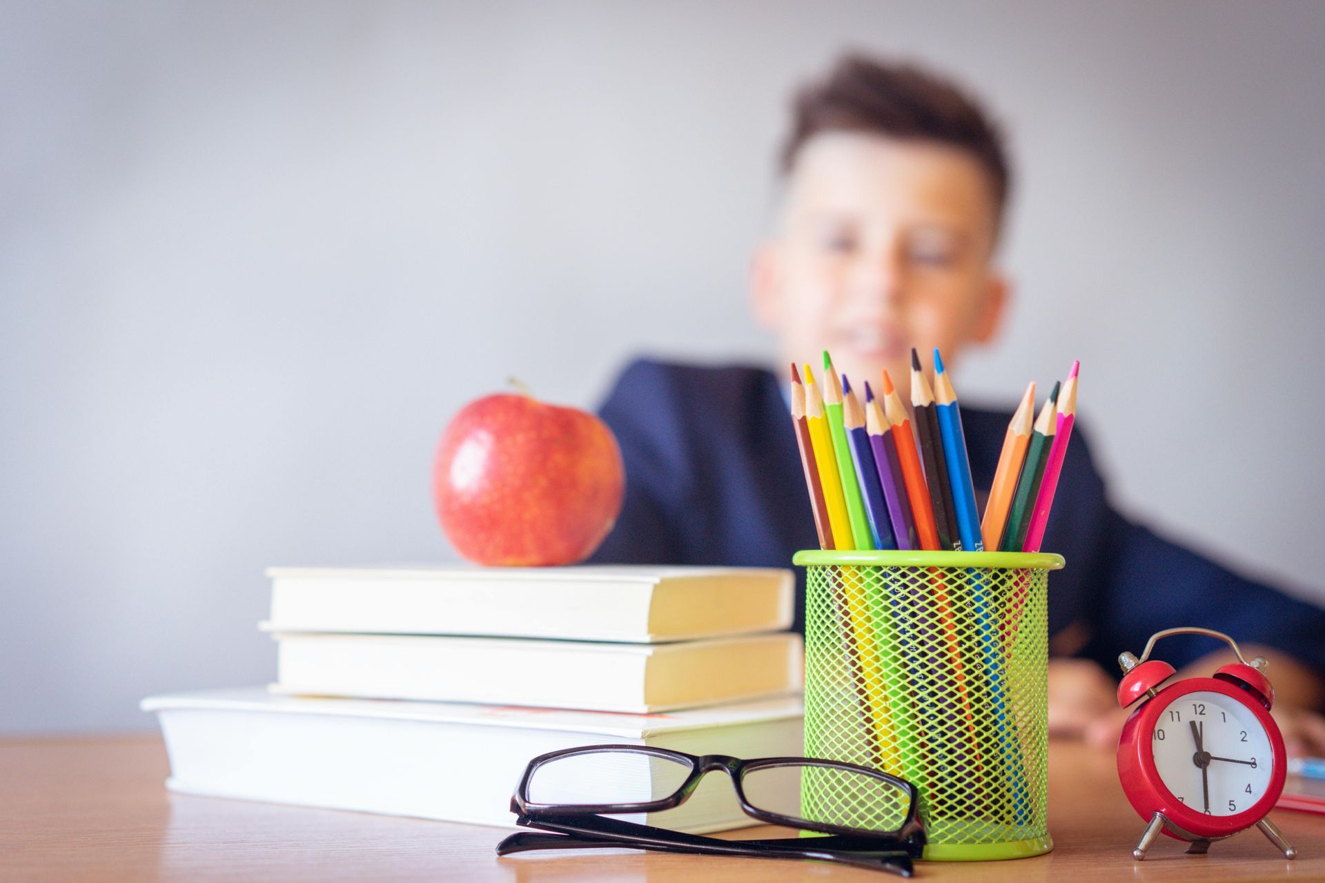 Desk with books, apple, pencils, glasses, and a clock; child in the background.