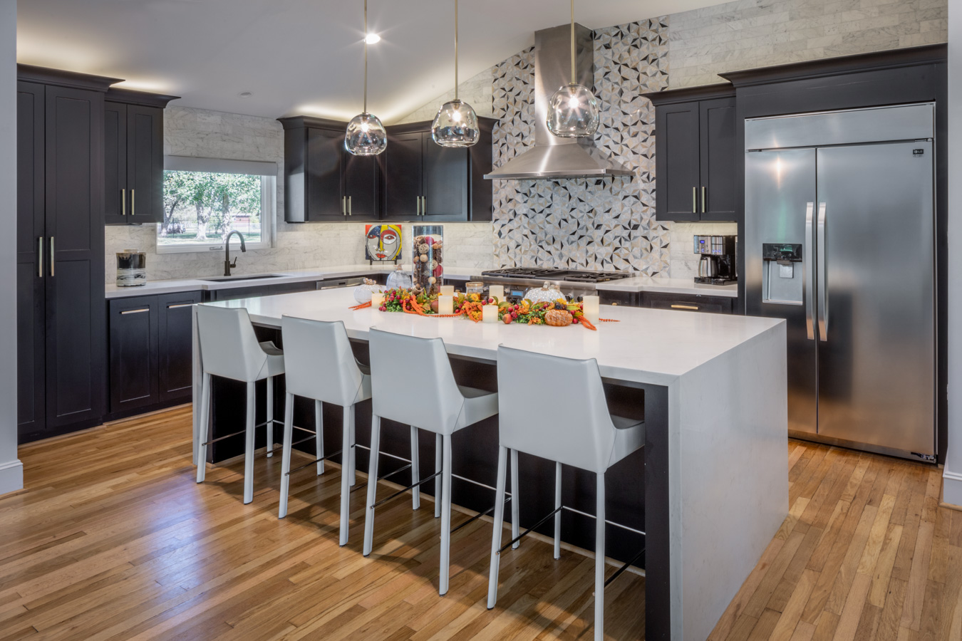 A kitchen with black cabinets , stainless steel appliances , a large island , and white chairs.