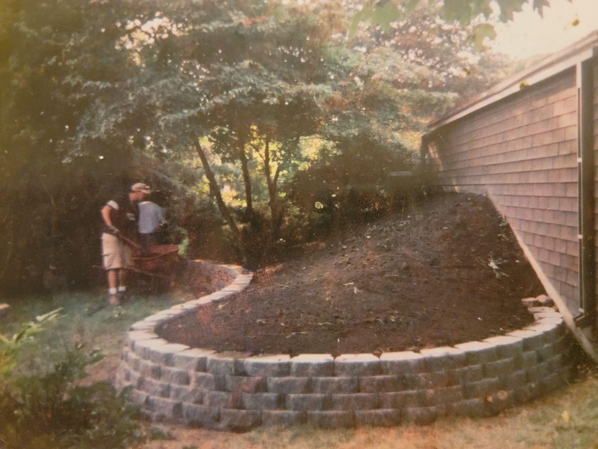 A man is pushing a wheelbarrow down a hill next to a brick retaining wall.