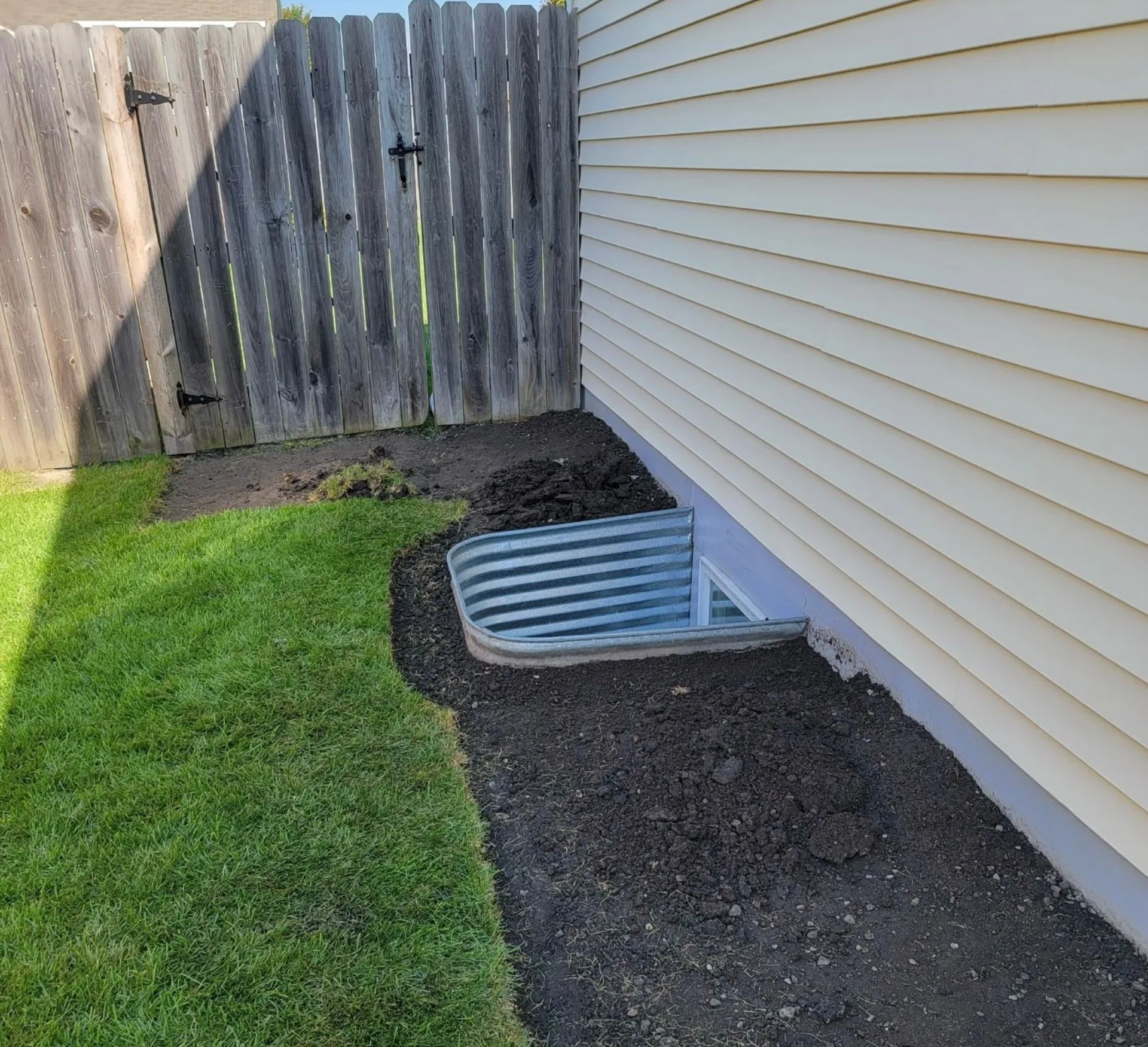 A window well in the side of a house next to a wooden fence.