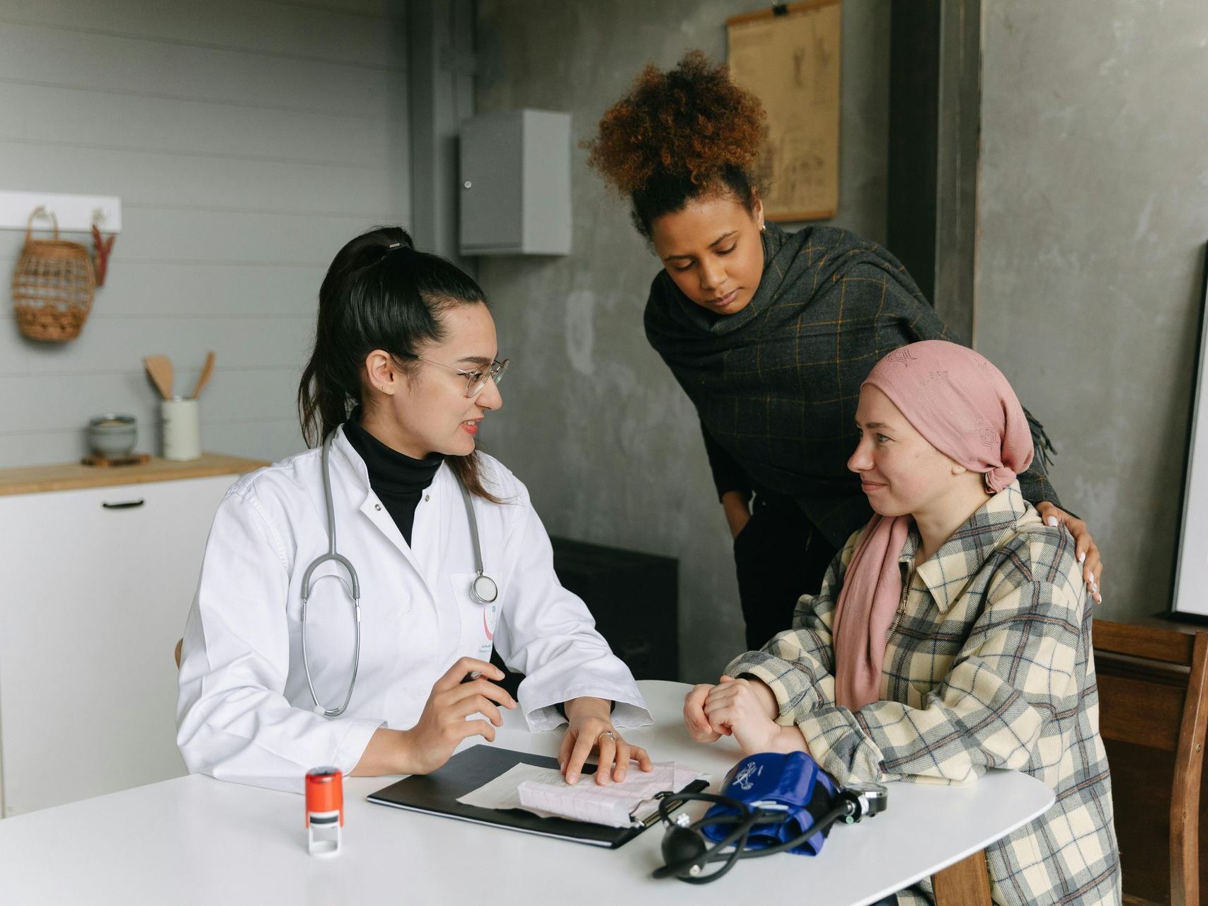 Health provider sitting with a woman with a scarf on her head at a table with another woman standing