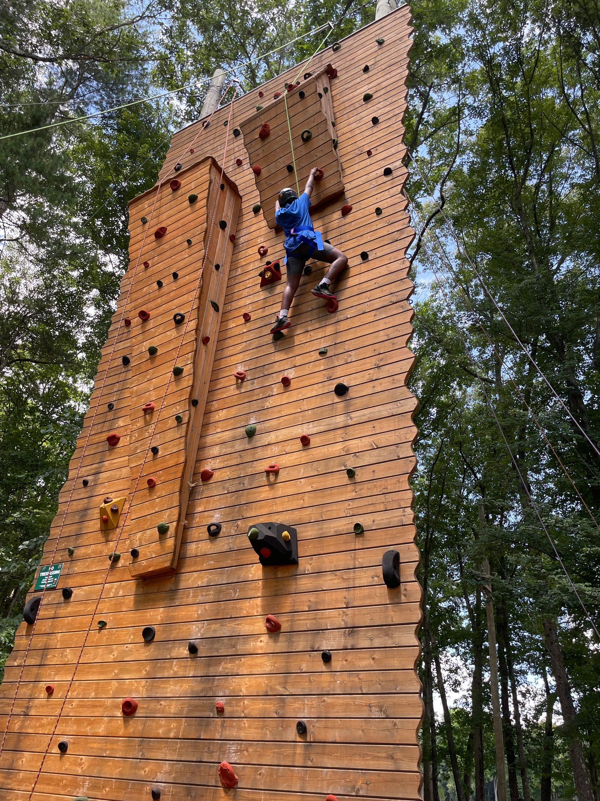 A man is climbing a wooden climbing wall in the woods.