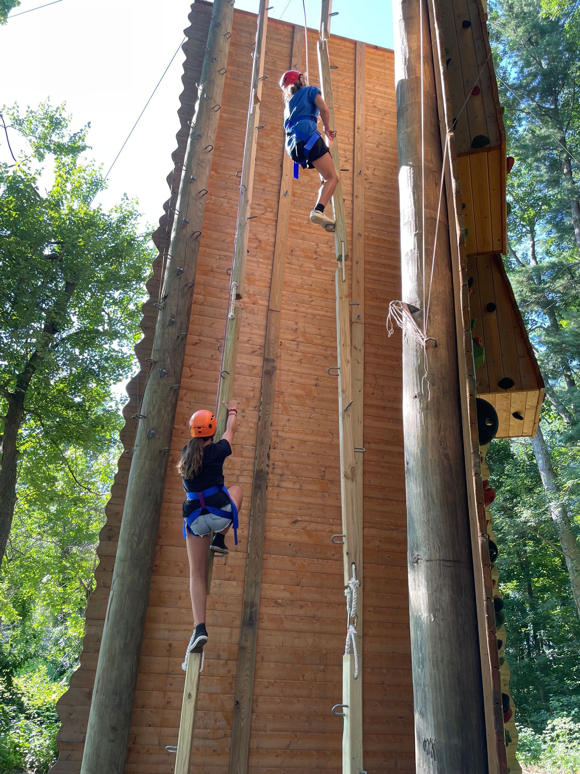 Two people are climbing up a wooden climbing wall.