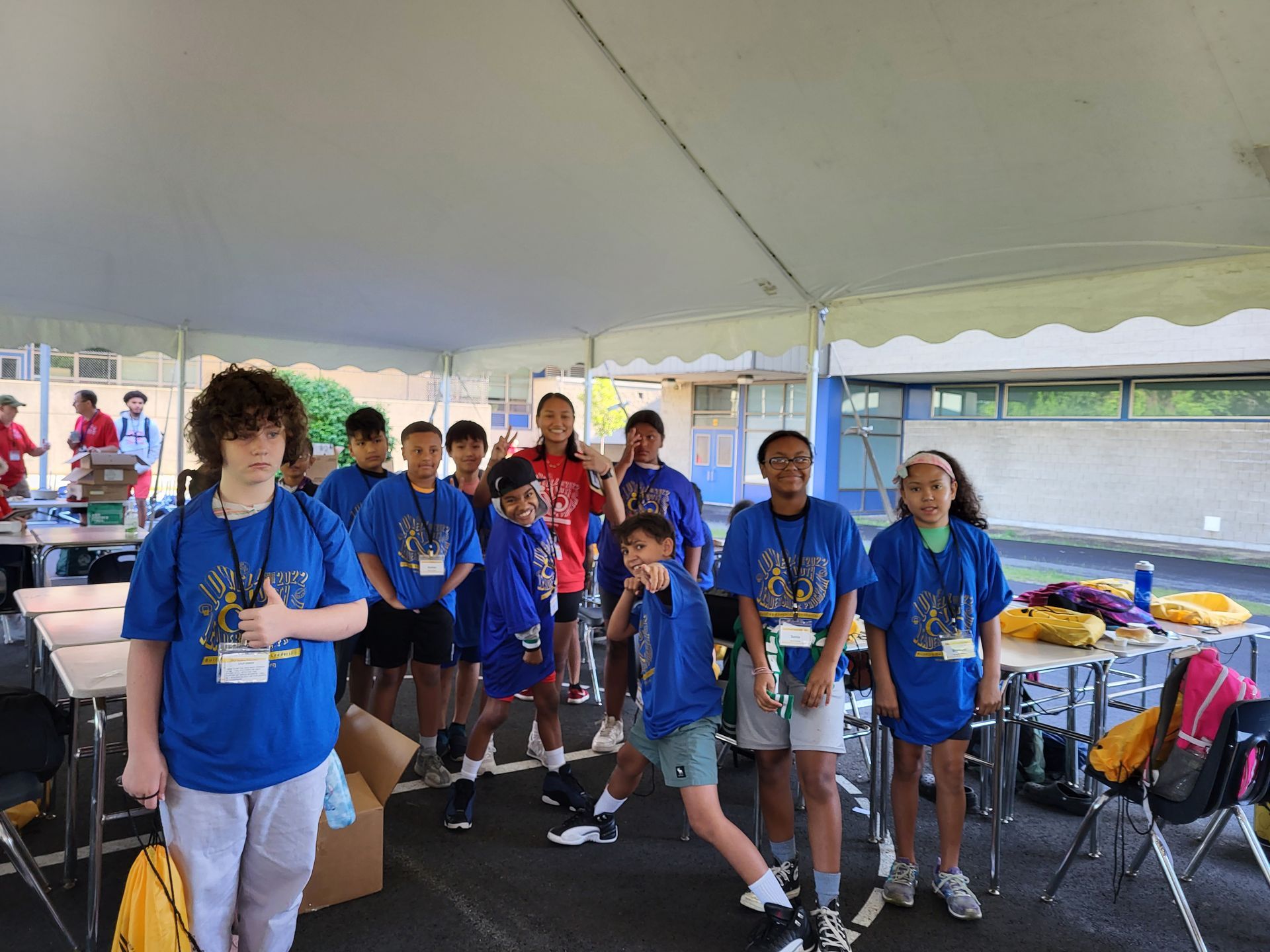 A group of children in blue shirts are standing under a tent.