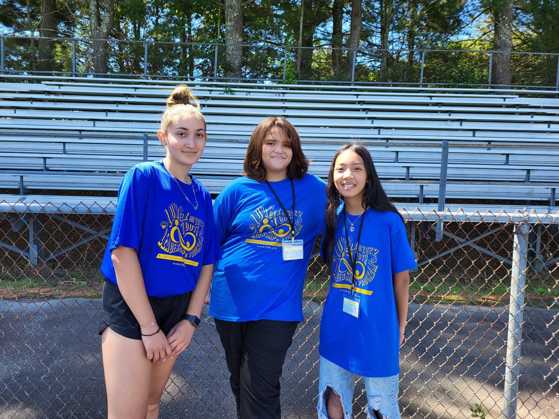 Three girls wearing blue shirts are posing for a picture in front of a bleacher.