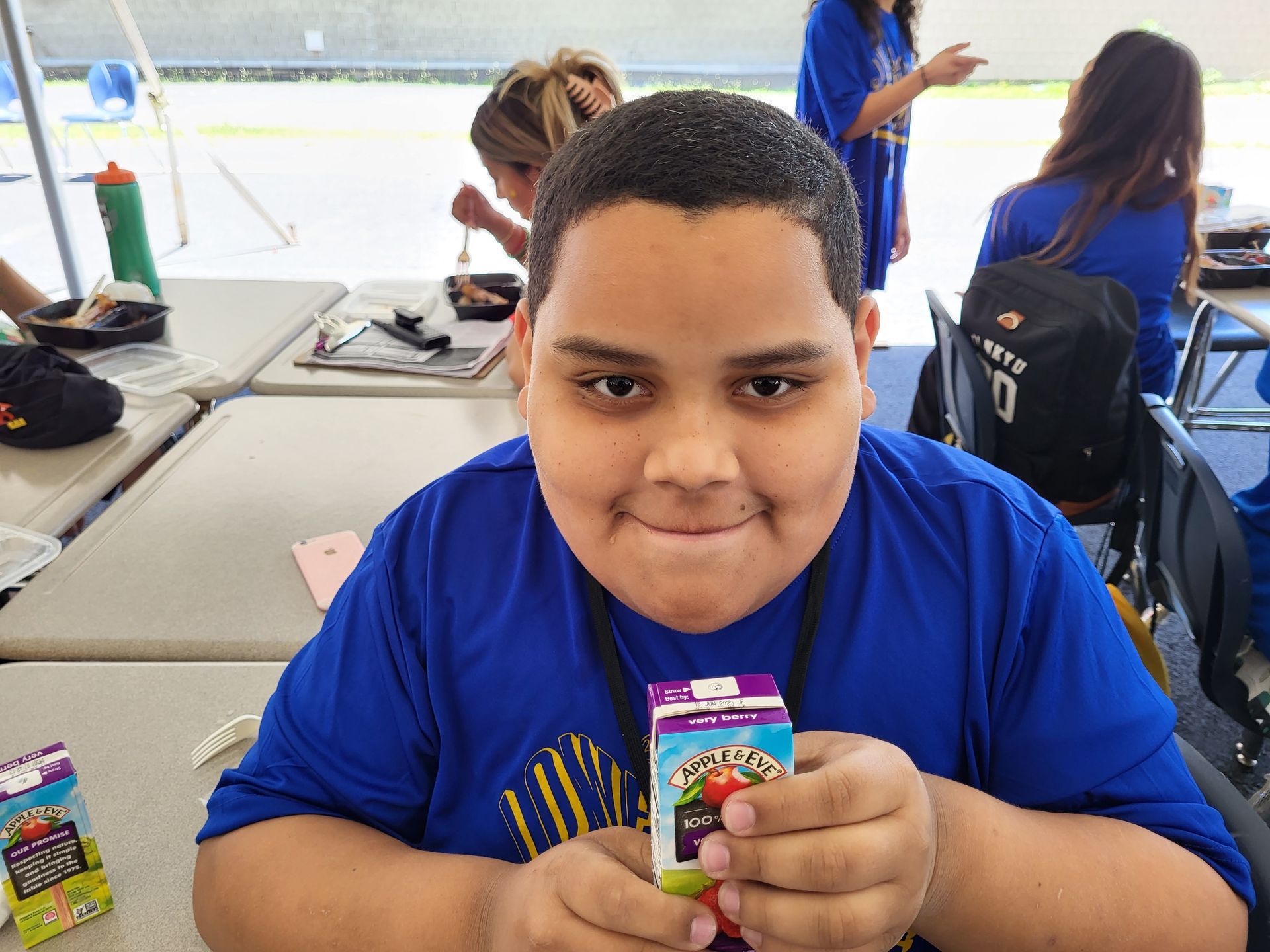 A boy in a blue shirt is sitting at a table holding a box of grape juice.