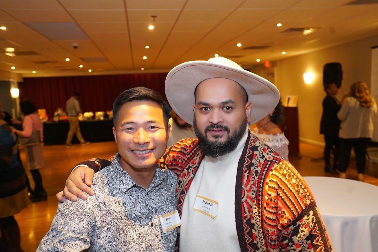 Two men are posing for a picture together in a room . one of the men is wearing a hat.