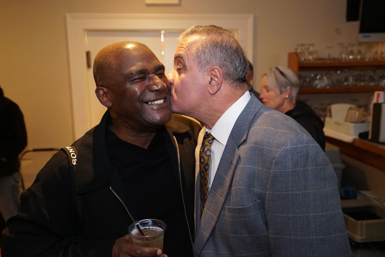 Two men are standing next to each other and one of them is kissing the other on the cheek.