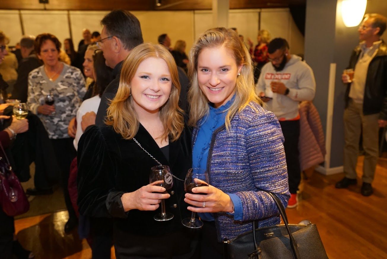 Two women are posing for a picture while holding wine glasses in a crowded room.