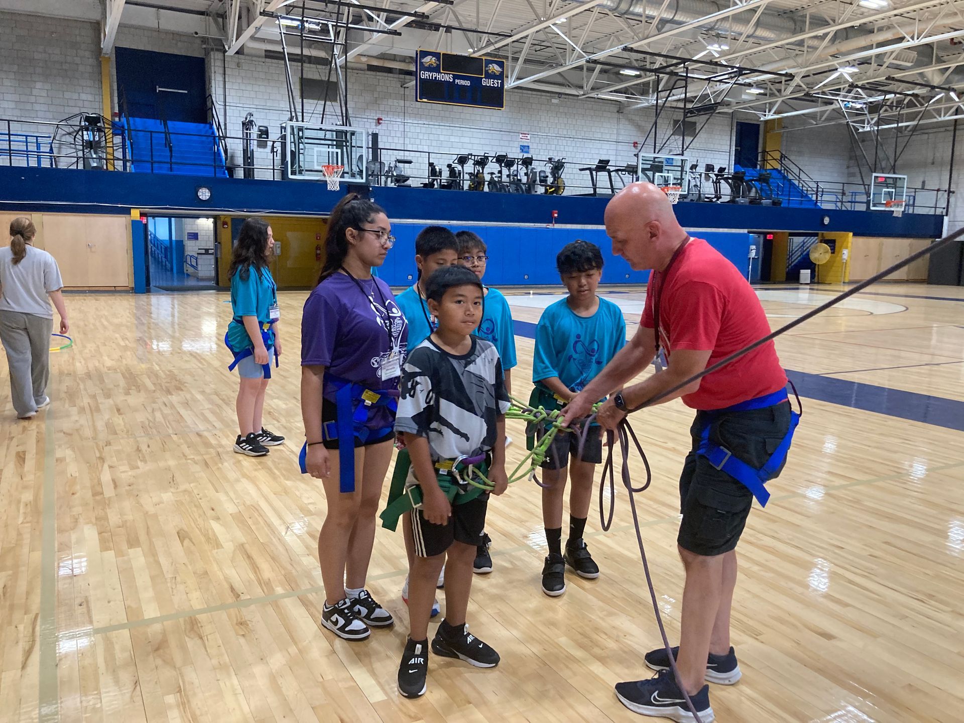 A man is teaching a group of children how to climb a rope in a gym.