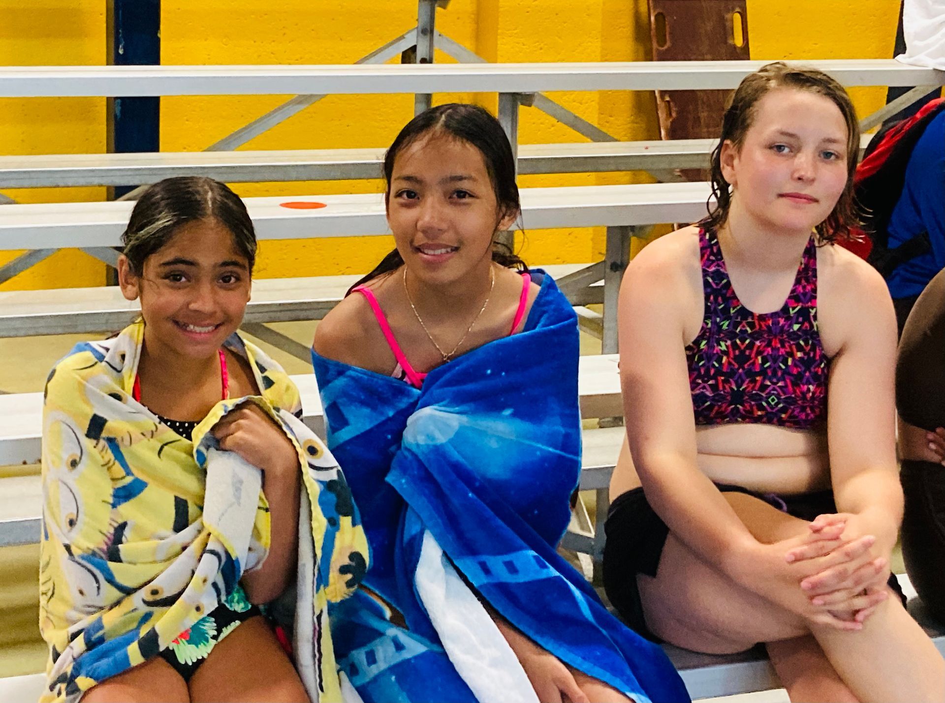 Three young girls wrapped in towels are sitting on bleachers at a pool.