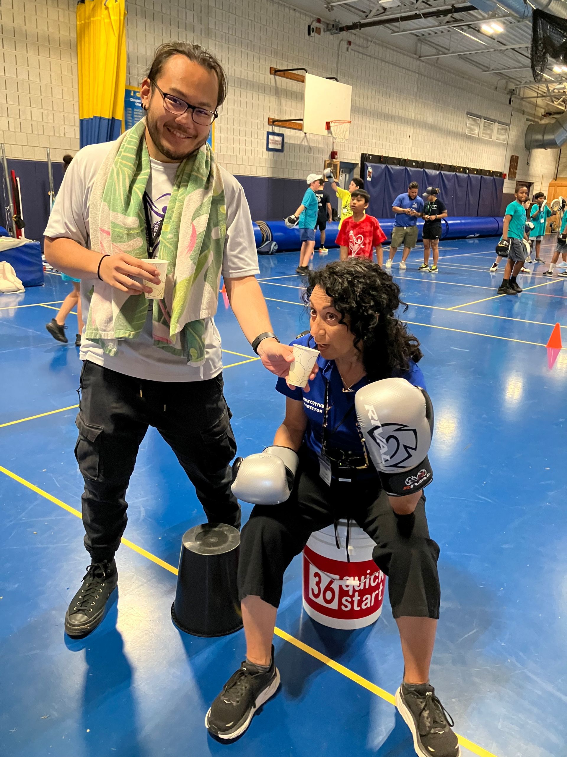 A man is standing next to a woman wearing boxing gloves in a gym.