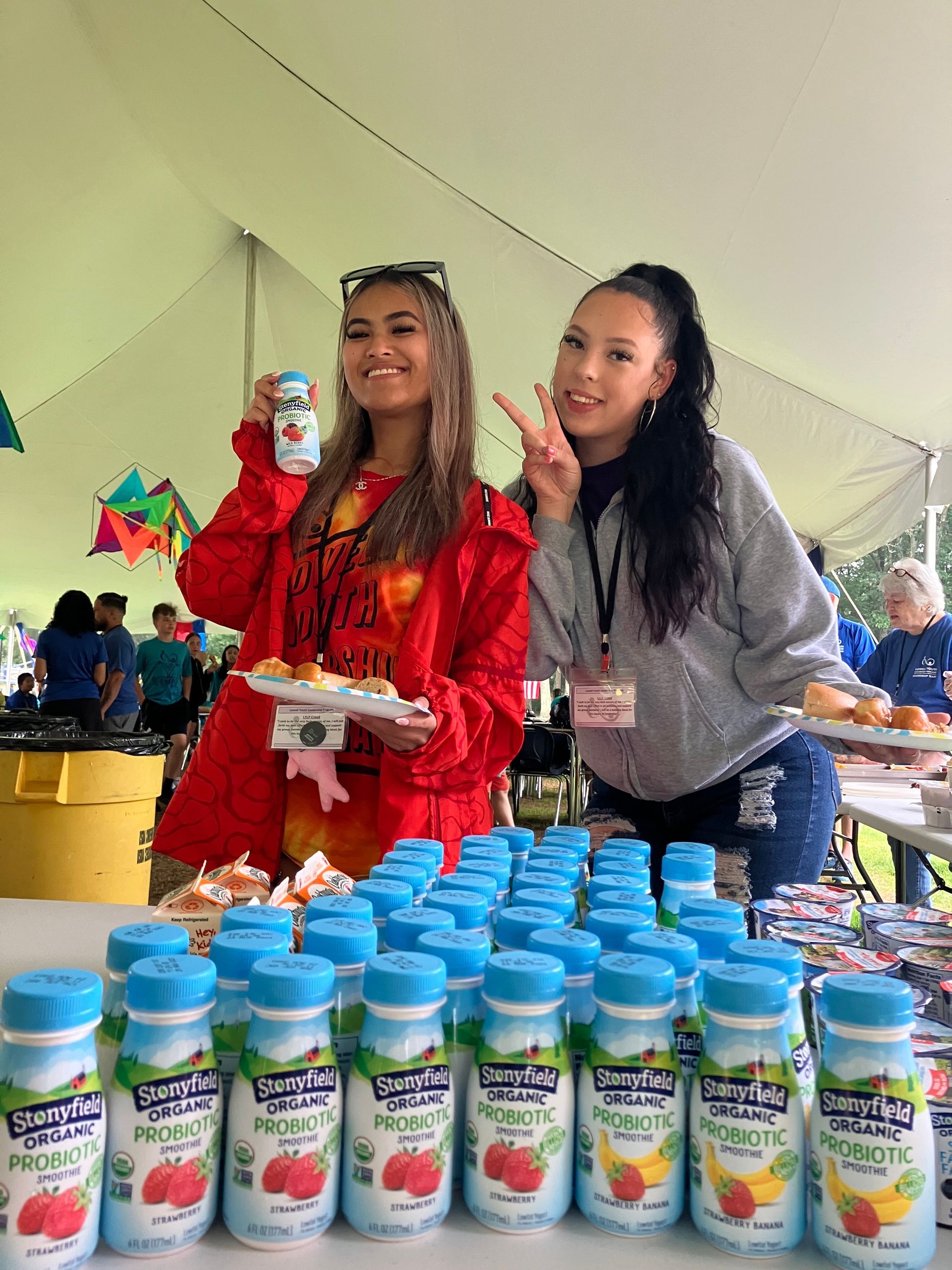 Two women are standing next to each other in front of a table filled with bottles of yogurt.