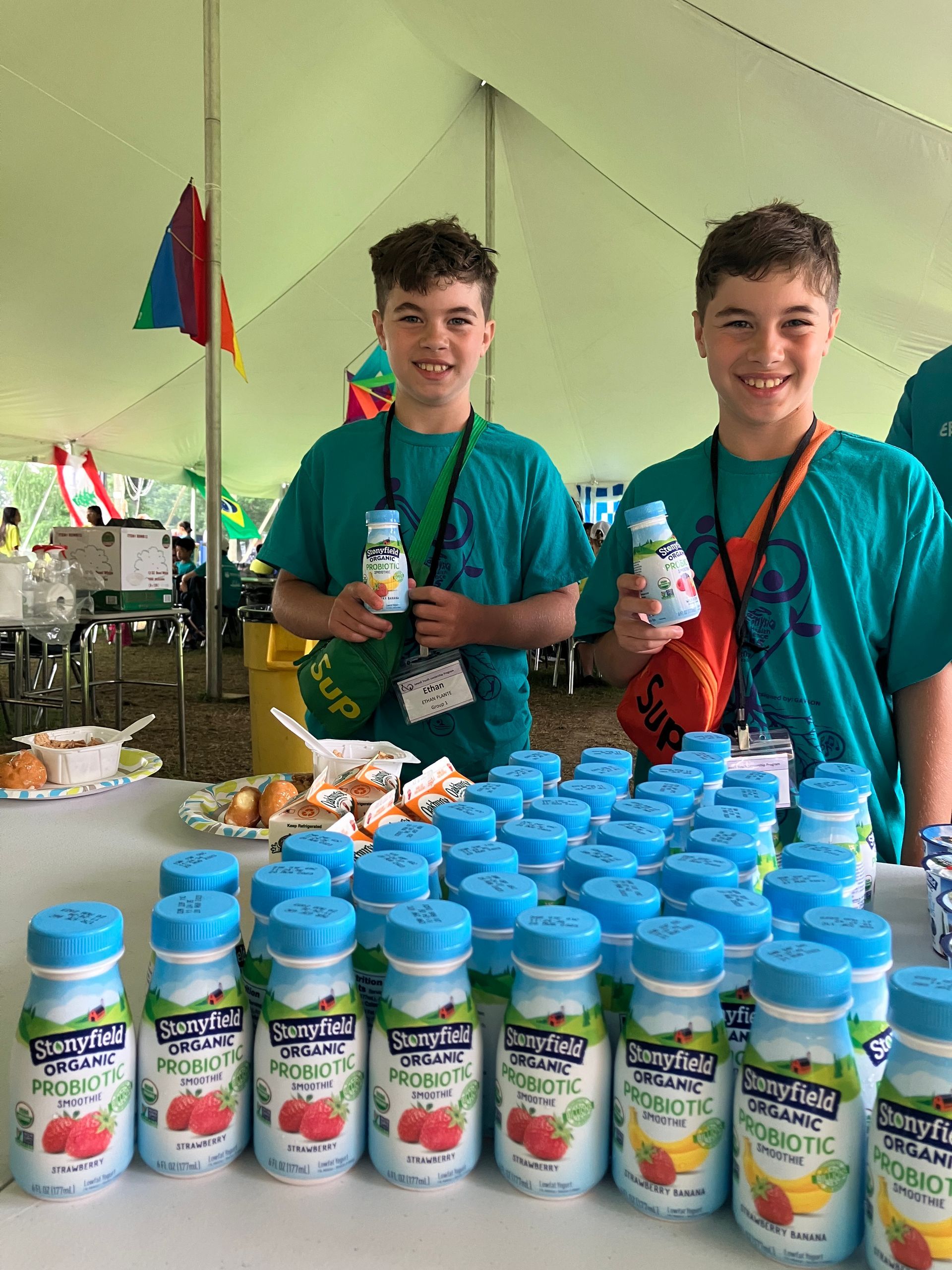 Two boys are standing in front of a table full of bottles of yogurt.