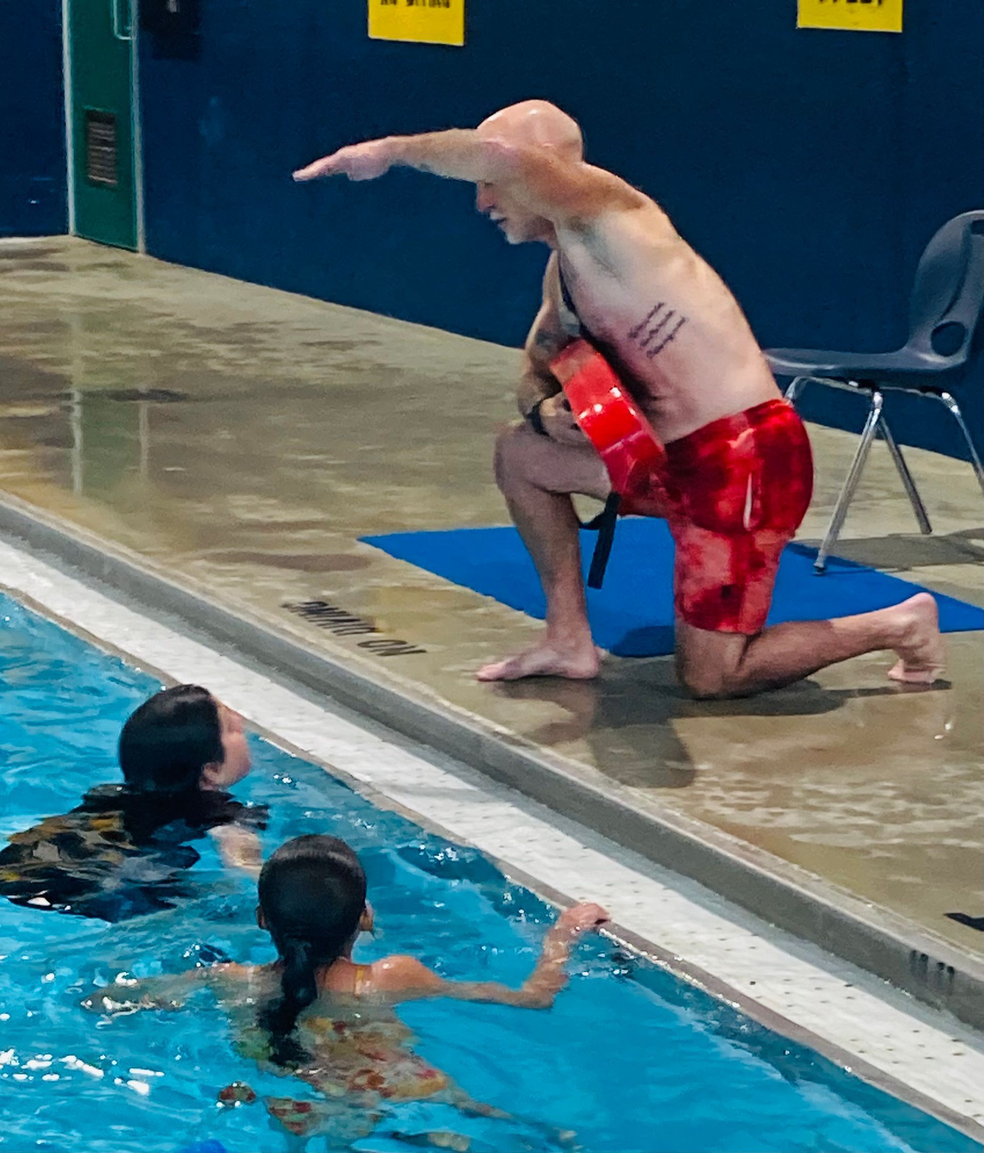 A man in red shorts is kneeling on the edge of a swimming pool