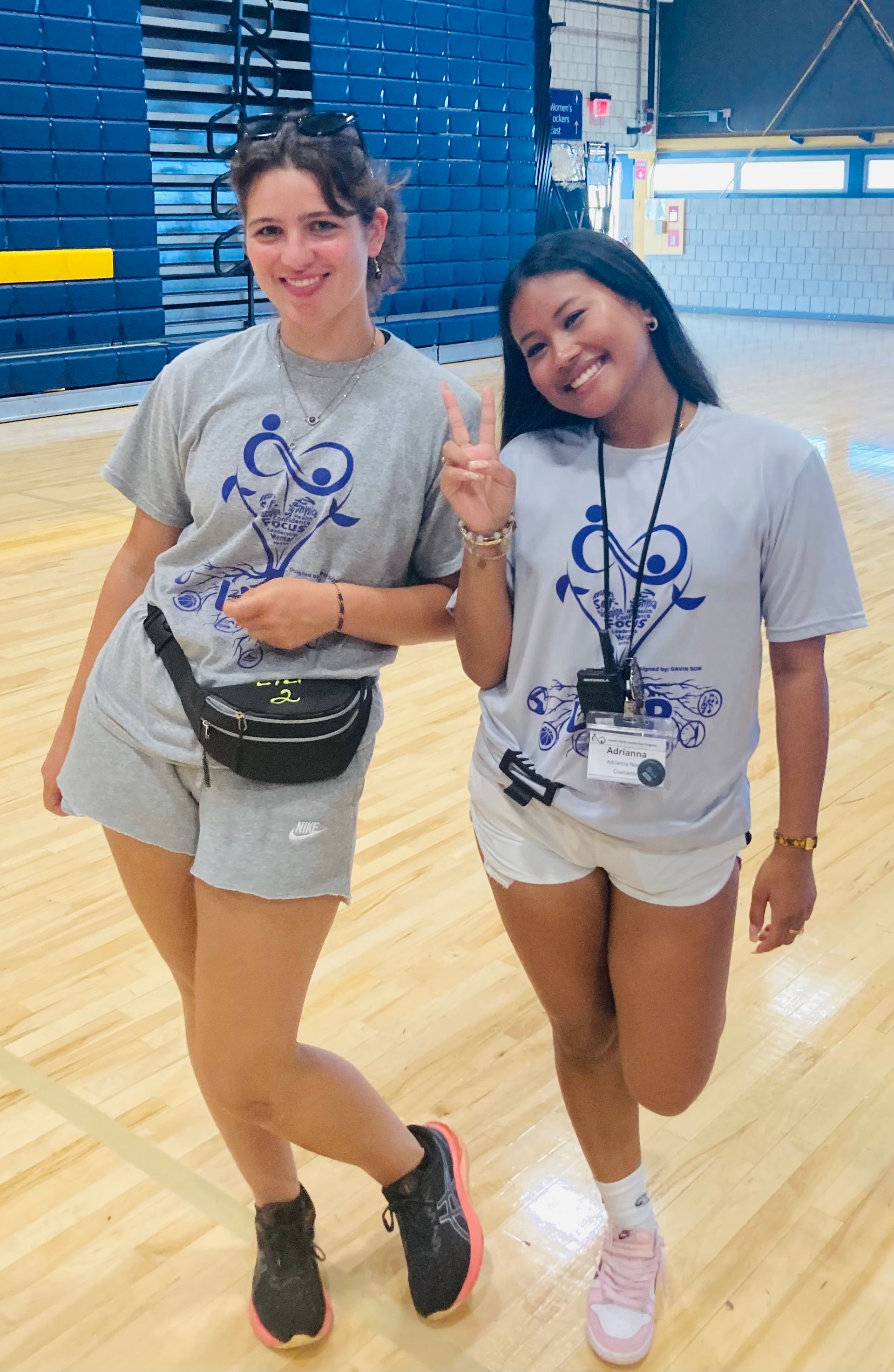 Two women are standing next to each other on a wooden floor in a gym.