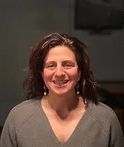 A woman wearing a grey sweater and earrings is smiling for the camera.