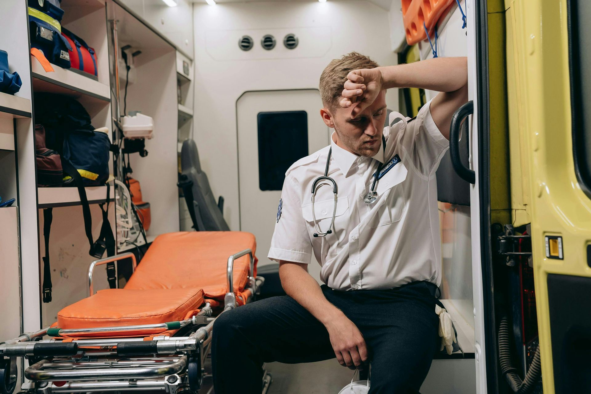 A paramedic with a stethoscope sits exhausted in the back of an ambulance, resting their head on their hand.