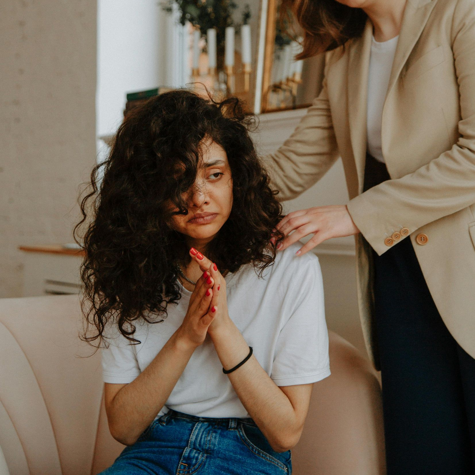 A distressed person in a white t-shirt and jeans is consoled by another person in a beige blazer, who rests a hand on them.