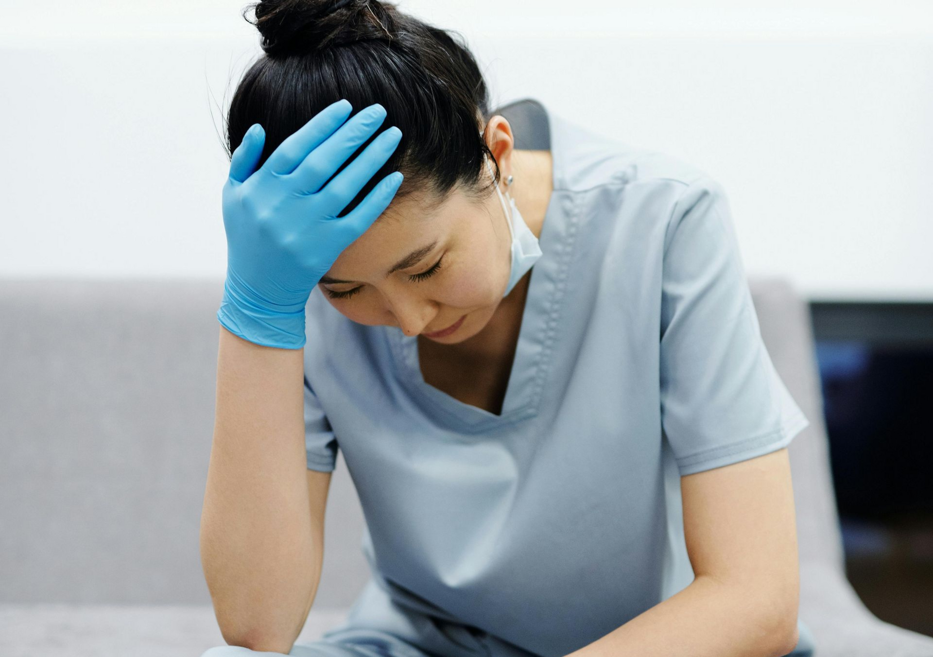 A healthcare worker in blue scrubs and a blue glove holds their head in exhaustion while sitting on a sofa.