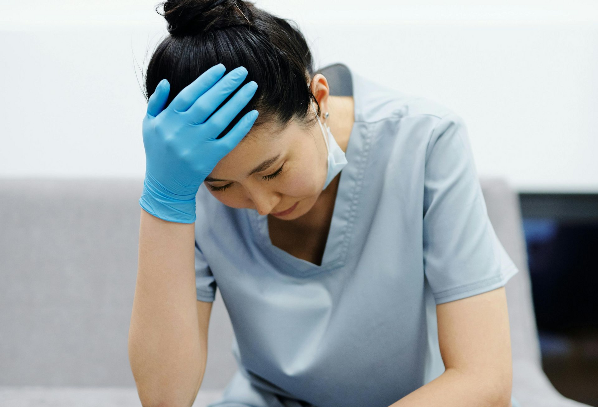 A medical professional in blue scrubs and gloves sits with their head in their hand, appearing distressed.