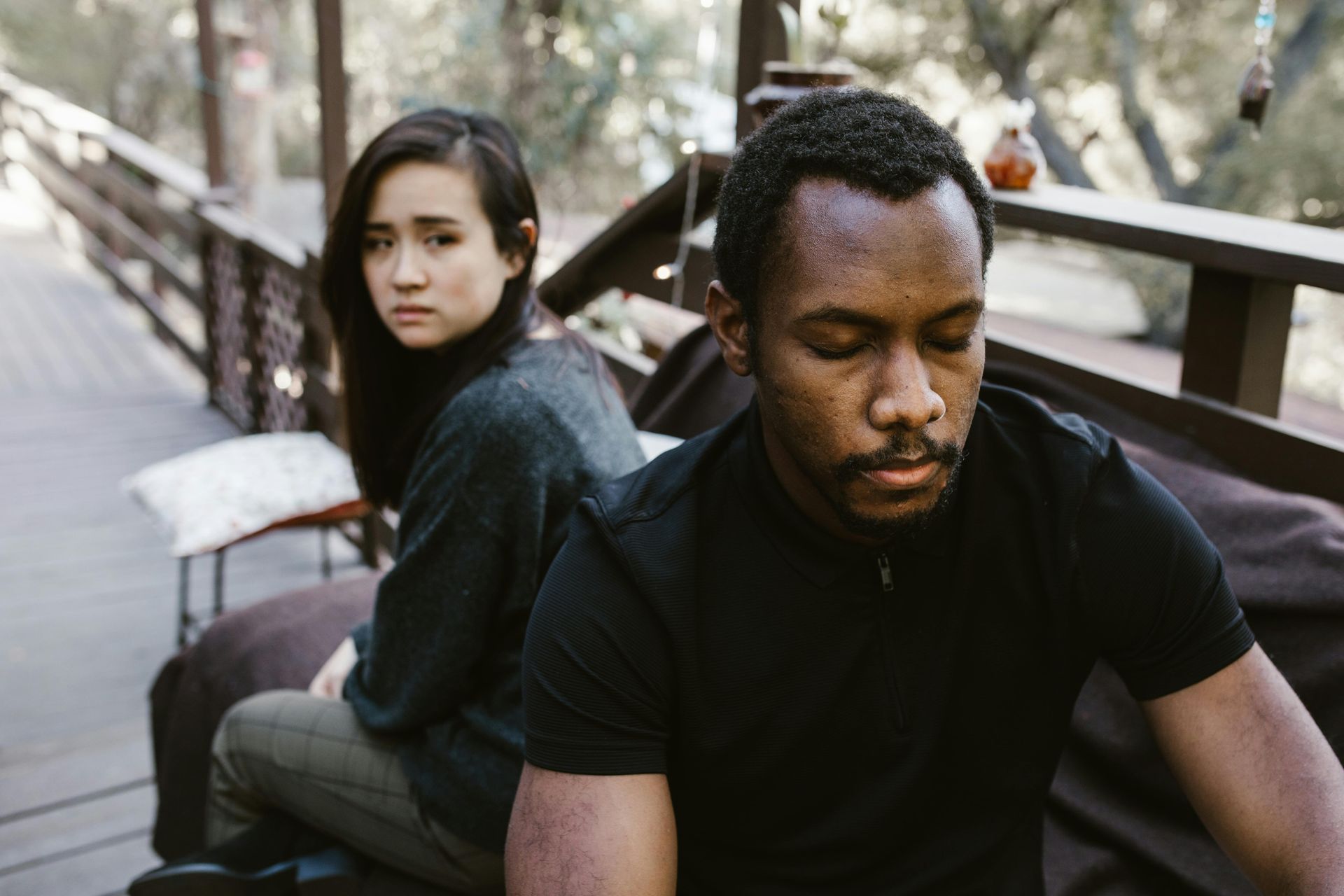 Two people sitting back-to-back on a wooden deck outdoors, appearing somber and distant.