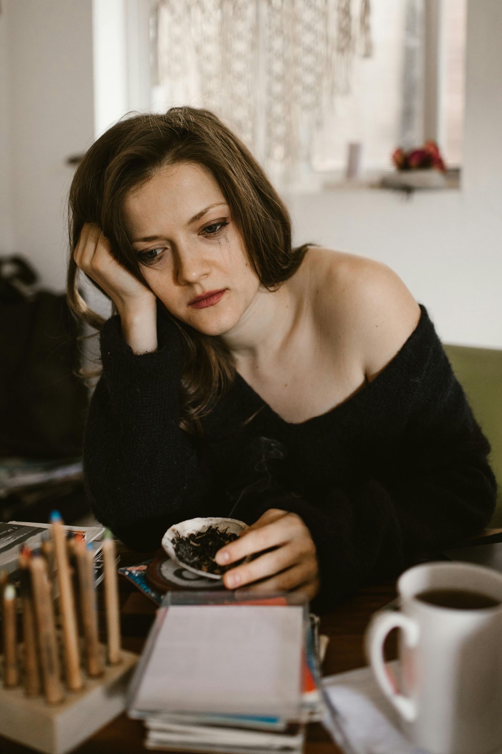 A person leans their head on their hand, looking down at a small dish while sitting at a desk with pencils and a mug.