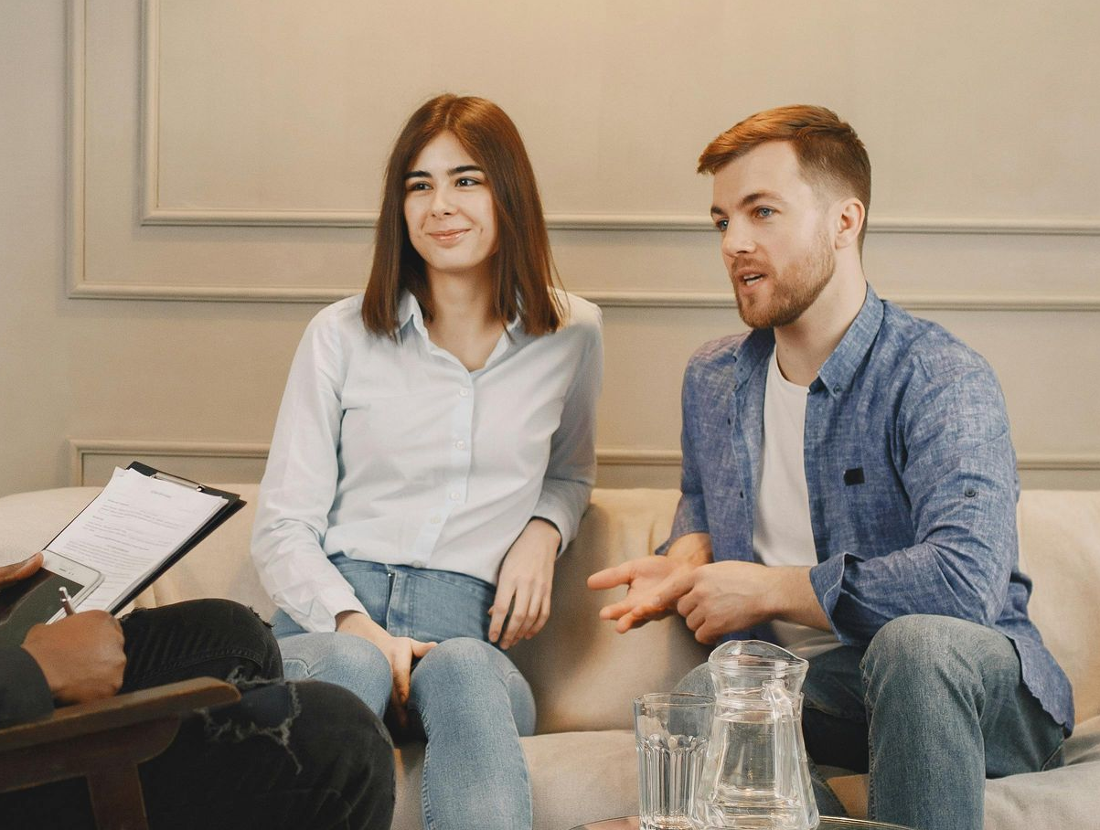 A couple talks to a professional during a session, sitting on a couch in a bright room with a desk in the foreground.