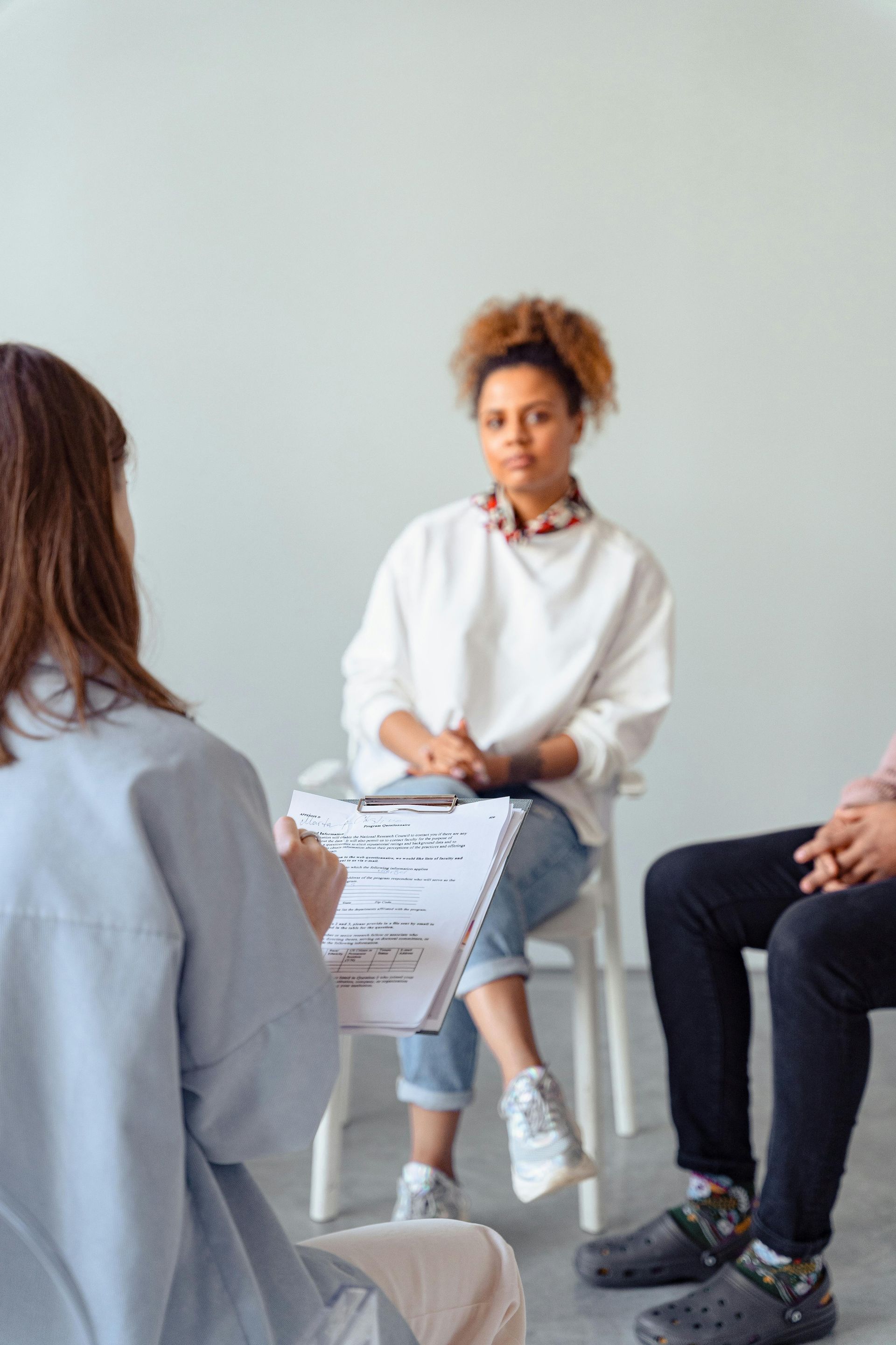 A therapist holds a clipboard while listening to two people seated in a group session against a plain wall.