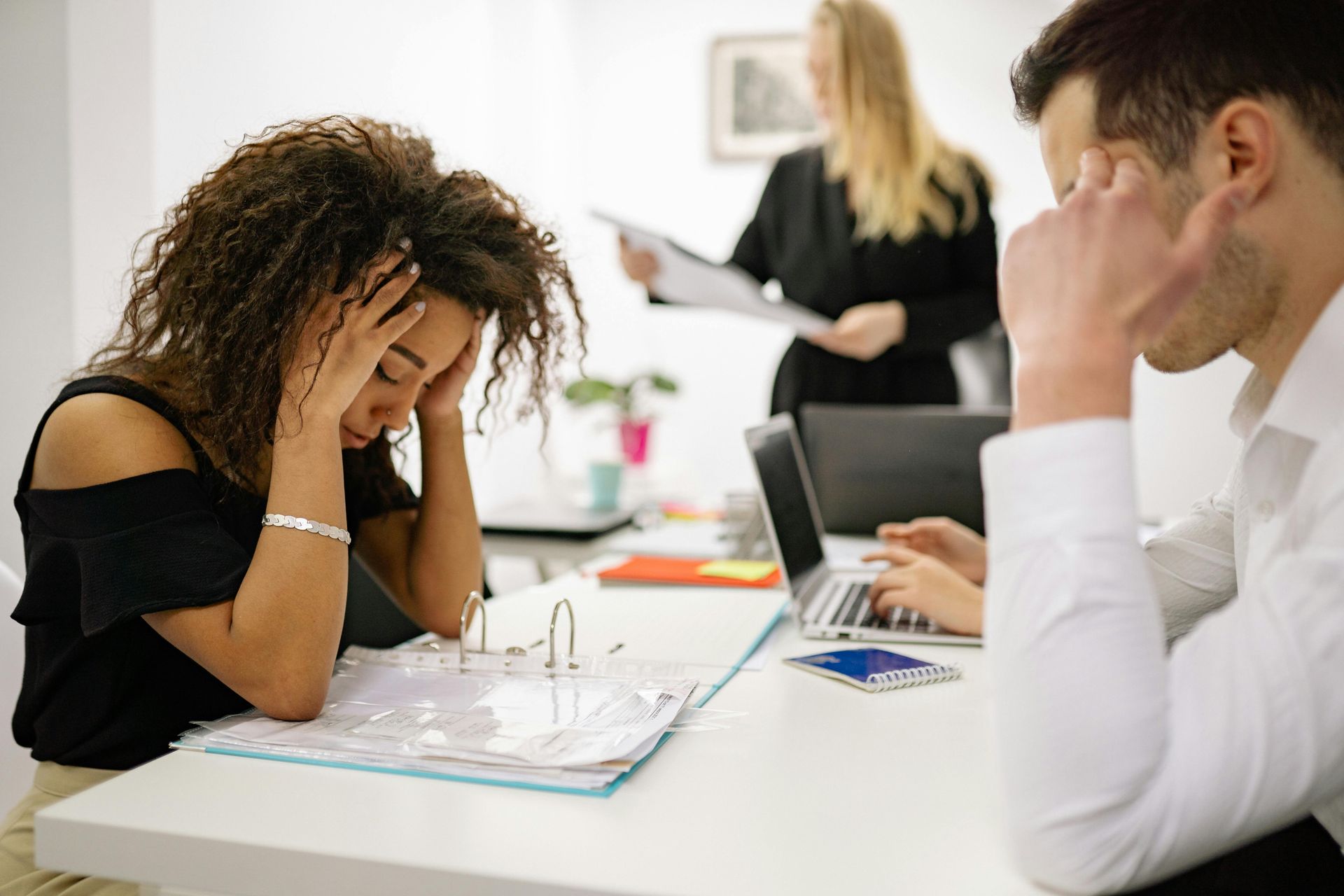 Two office workers sit at a desk, looking stressed with hands on their heads, while a colleague works in the background.