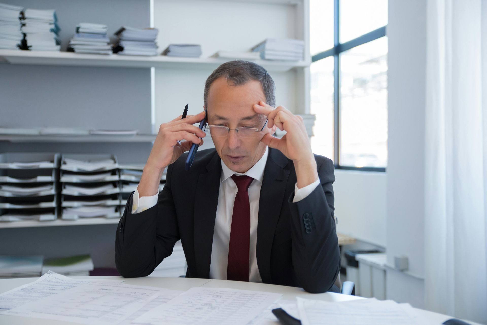 A person in a suit sits at a desk with papers, looking stressed with hands on their temples while holding a pen.