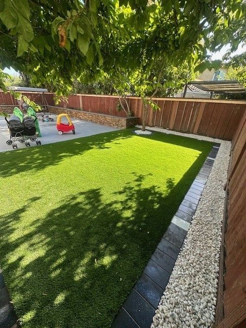A green lawn bordered by rocks and pavers, with a wooden fence and tree, with kids in the background.
