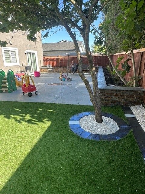 Backyard with artificial grass, a tree surrounded by white stones, and children playing on a concrete patio.