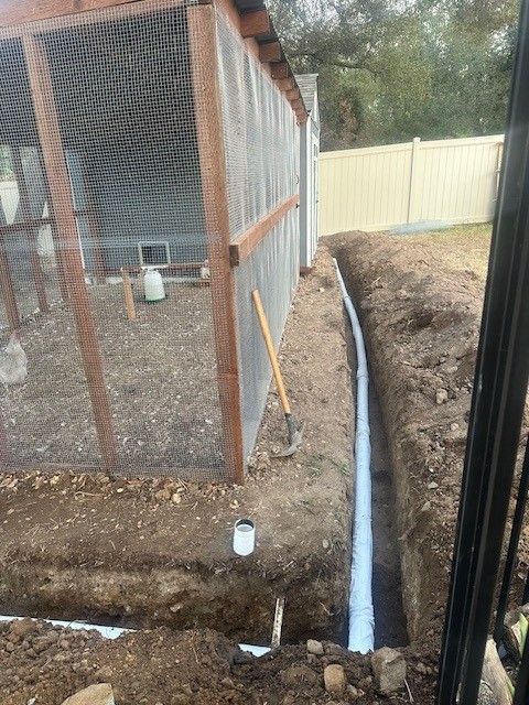 Trench dug next to a chicken coop for drainage pipes. Soil, white fence and wood frame visible.