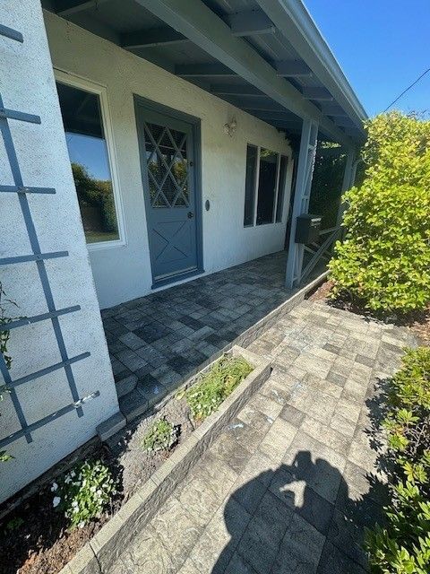 A narrow paved walkway leads to a blue door with a latticework design, under a covered porch.