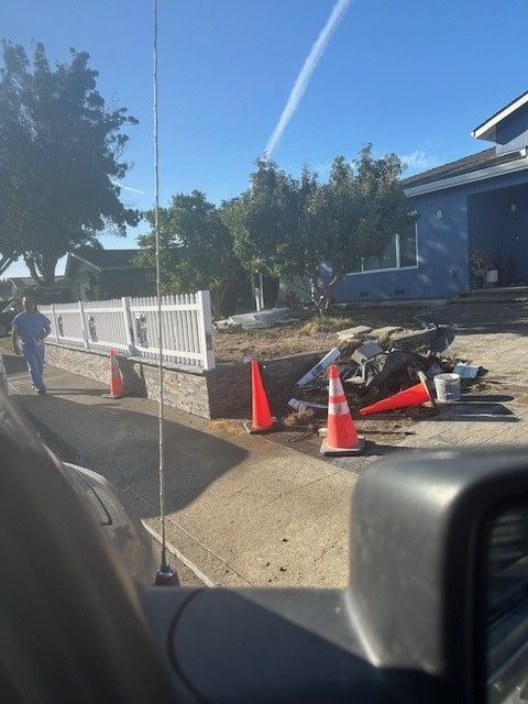 Construction scene: orange cones, brick wall, blue house, man walking on sidewalk.