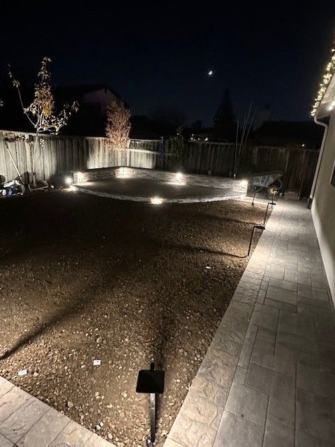 Backyard at night, illuminated by spotlights. Dirt yard, stone patio, wooden fence, and moon visible.