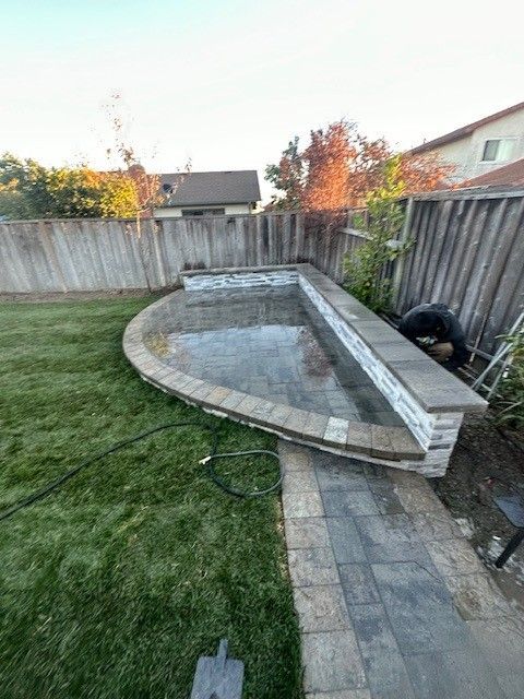Backyard with newly constructed patio, low wall, and walkway; green grass and wooden fence in background.