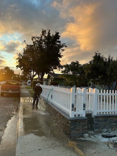Person power washing a white picket fence and sidewalk. Cloudy sunset sky and street with a truck.