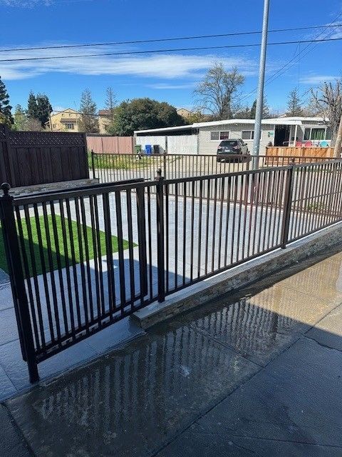 Black metal fence surrounding a paved area and grass. Building and sky in the background.