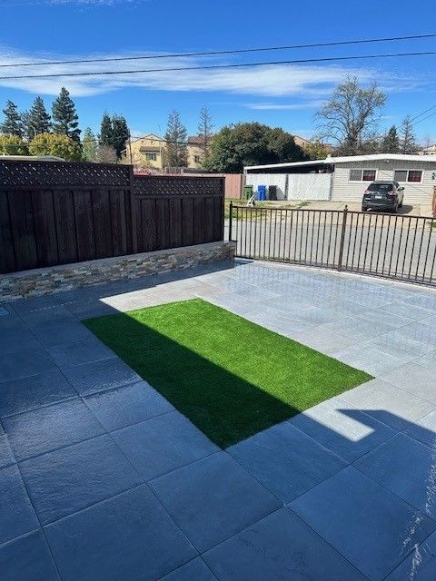 Backyard patio with gray tiles, dark wood fence, patch of green turf, and sunny blue sky.