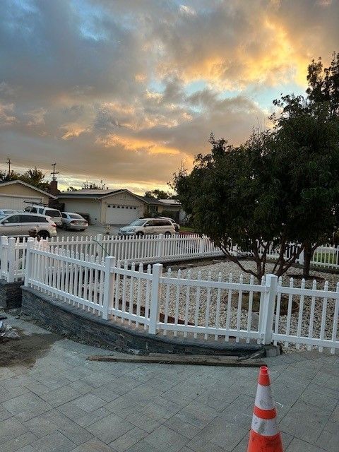 White picket fence surrounds a landscaped area with a tree. Sunset sky above houses and cars.