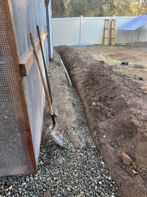 A trench dug along a building, with a shovel leaning against it; gravel on the bottom.