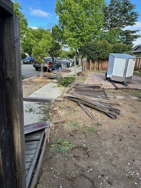 Debris and a trailer next to a sidewalk with broken wooden fence in a neighborhood.
