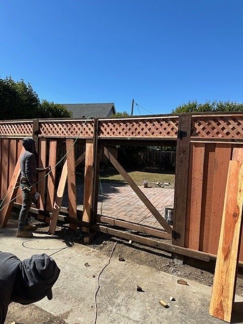 Two workers building a wooden gate in a backyard. One uses a tool. The sky is blue.