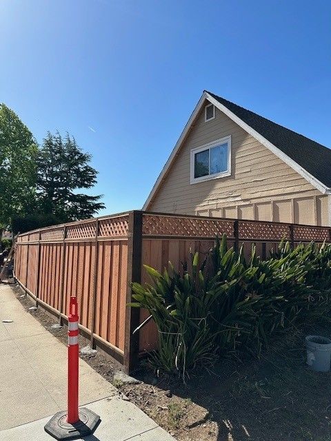 A wood fence with lattice-top surrounds a tan house. A red and white pole sits on the sidewalk.