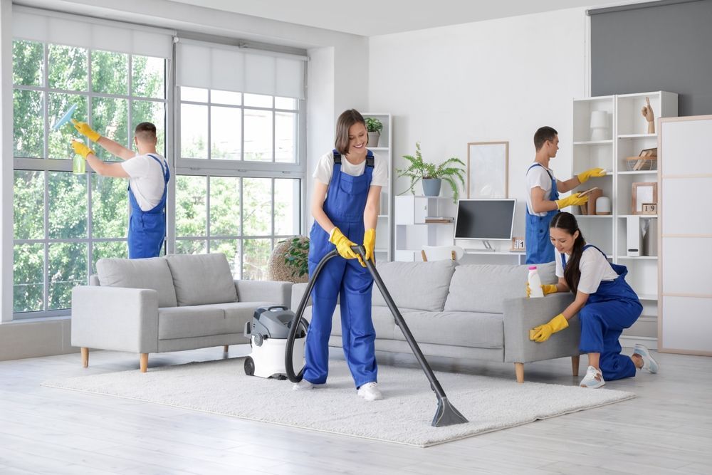 Cleaning crew in blue jumpsuits cleaning a living room with windows, sofas, and shelves.