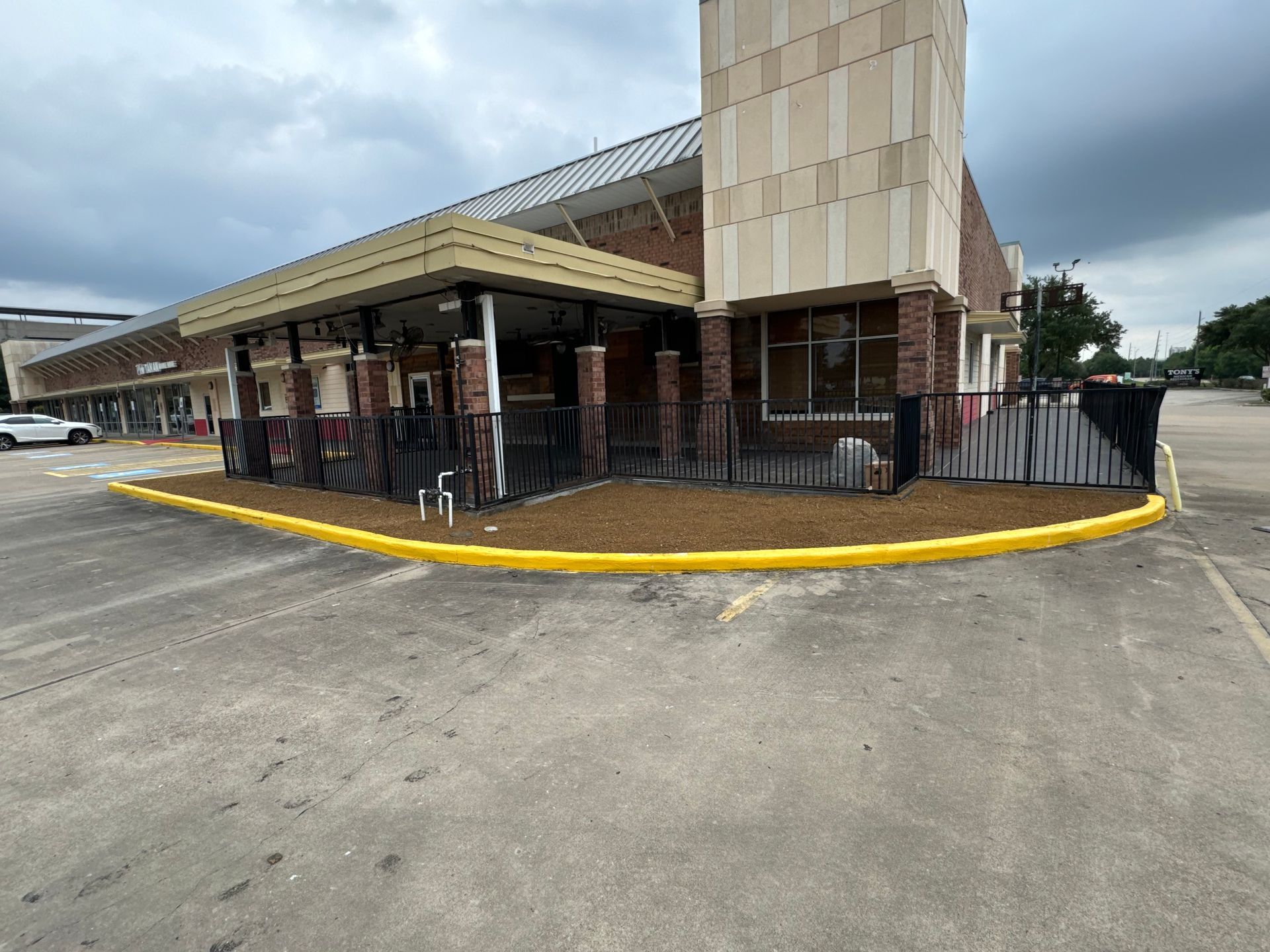 Exterior of a brick building with a covered outdoor area enclosed by a black fence, with brown mulch and a yellow curb.