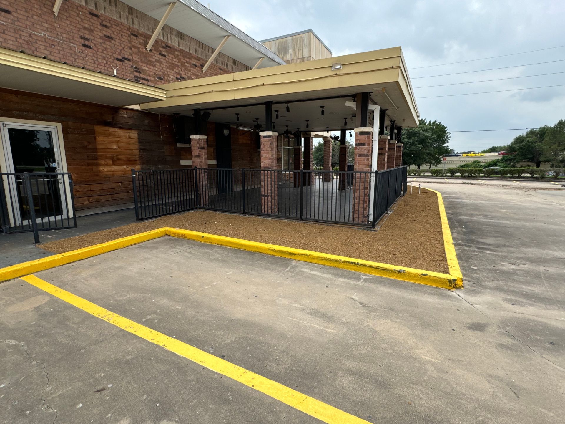 Exterior of a restaurant with a covered patio. Red brick building, gravel landscaping, black metal fence, yellow parking lines.